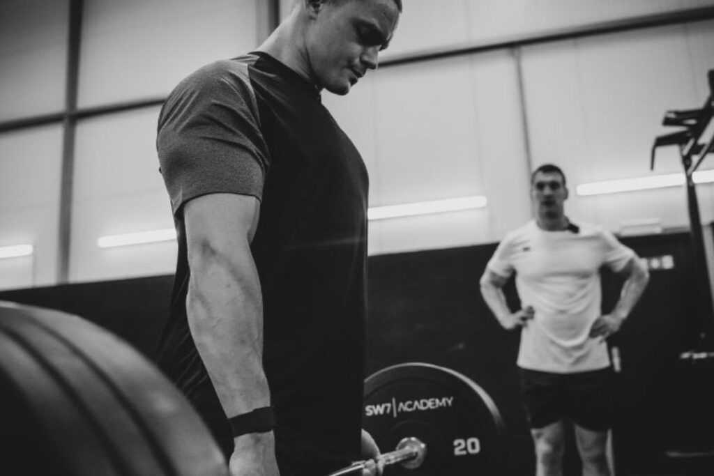 Man holding barbell and another man stood watching.