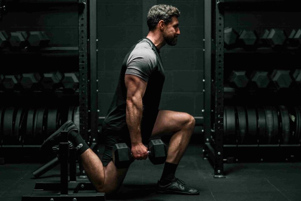 Josh doing a Bulgarian split squat at the gym, using a black split squat stand for his back leg, with racks of weights behind.