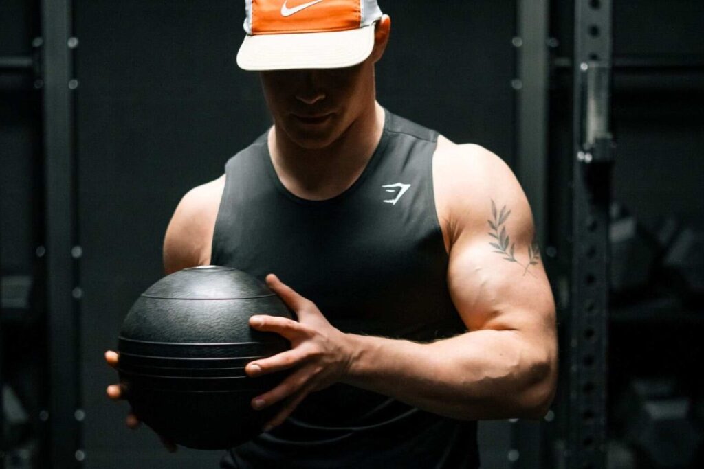 Gassy, wearing a baseball cap, holds a black medicine ball at the gym.
